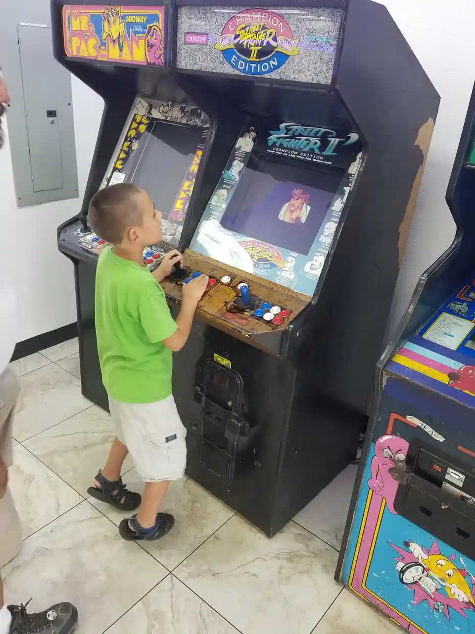 A young boy in a green shirt and white shorts is standing at a vintage Street Fighter II Champion Edition arcade machine…