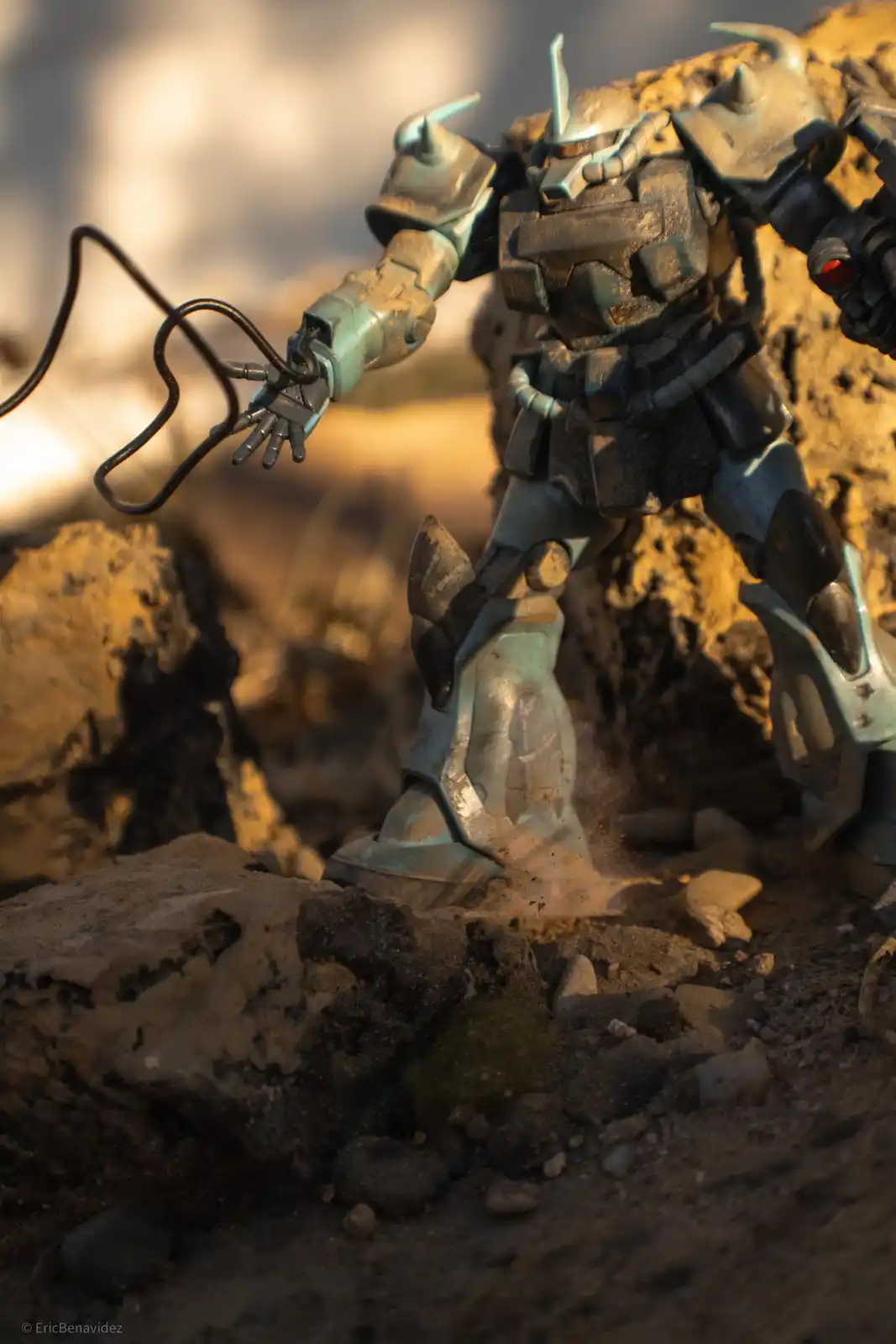 A weathered, teal-colored robot action figure stands on rocky terrain, its right hand gripping a black electrical cord that…