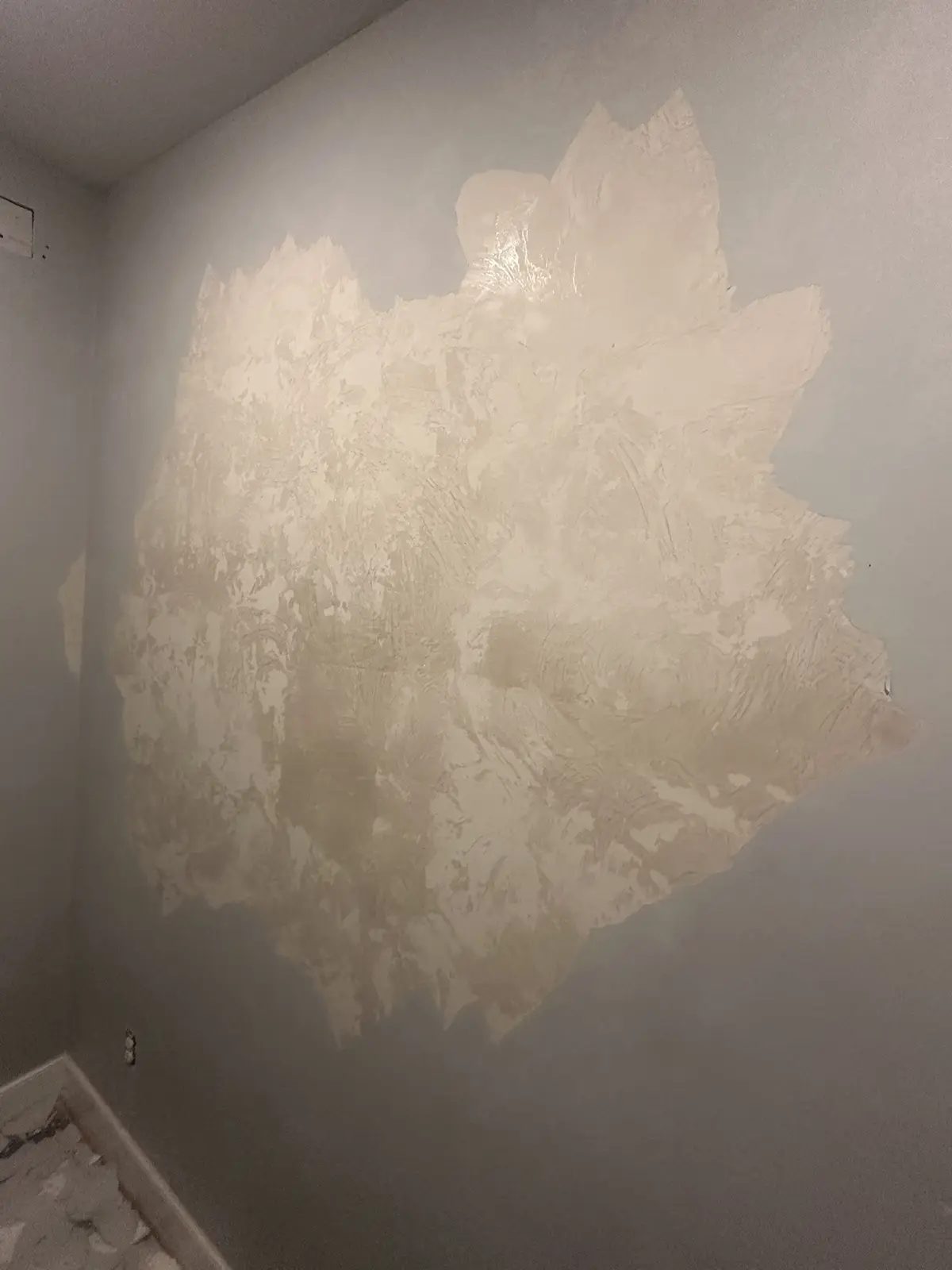 A close-up photo shows a large, irregularly shaped patch of beige drywall mud applied to a gray wall in a corner, with…