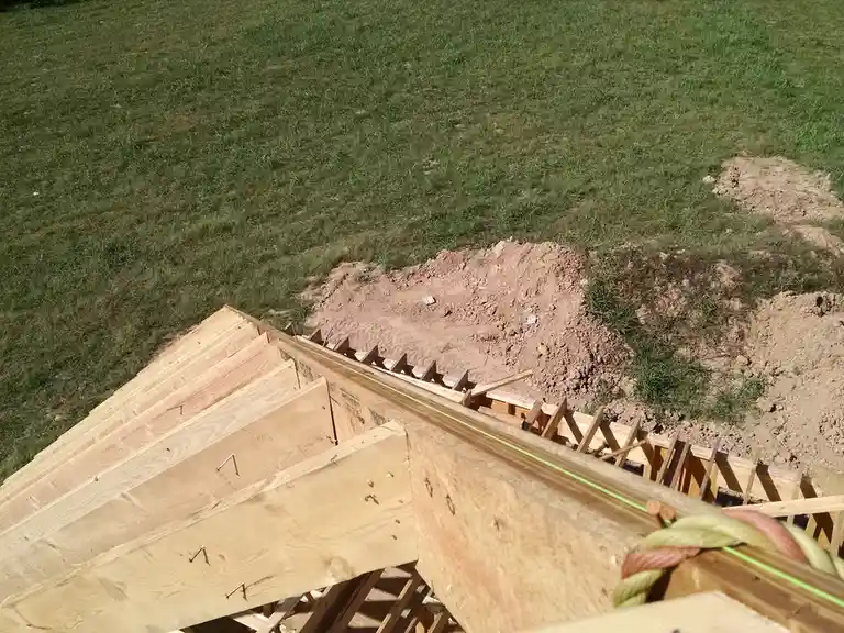 An overhead view of a partially constructed wooden house frame on a grassy lot, with dirt piles nearby. A green laser line…