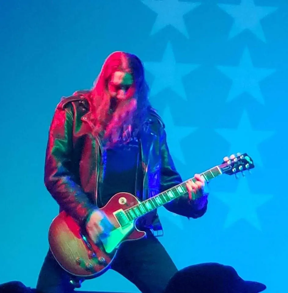 A musician with long hair and a leather jacket plays a red electric guitar on stage under blue lighting, with a backdrop of…