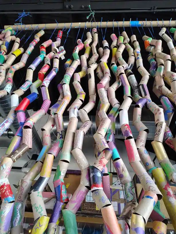 A colorful, chaotic art installation made from hundreds of decorated cardboard toilet paper rolls hanging from a wooden rod…