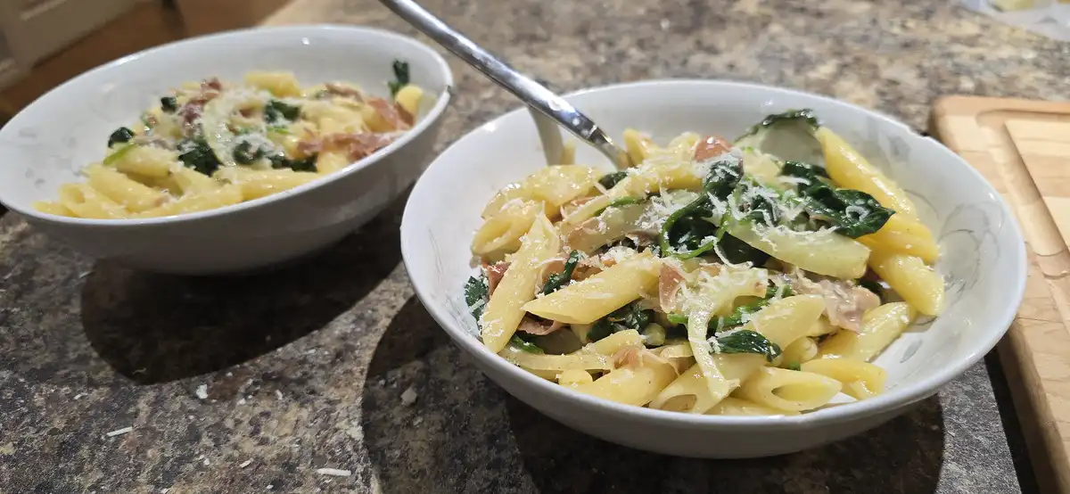 Two white bowls of penne pasta with spinach, bacon, and grated cheese sit on a granite countertop, one with a fork inserted…