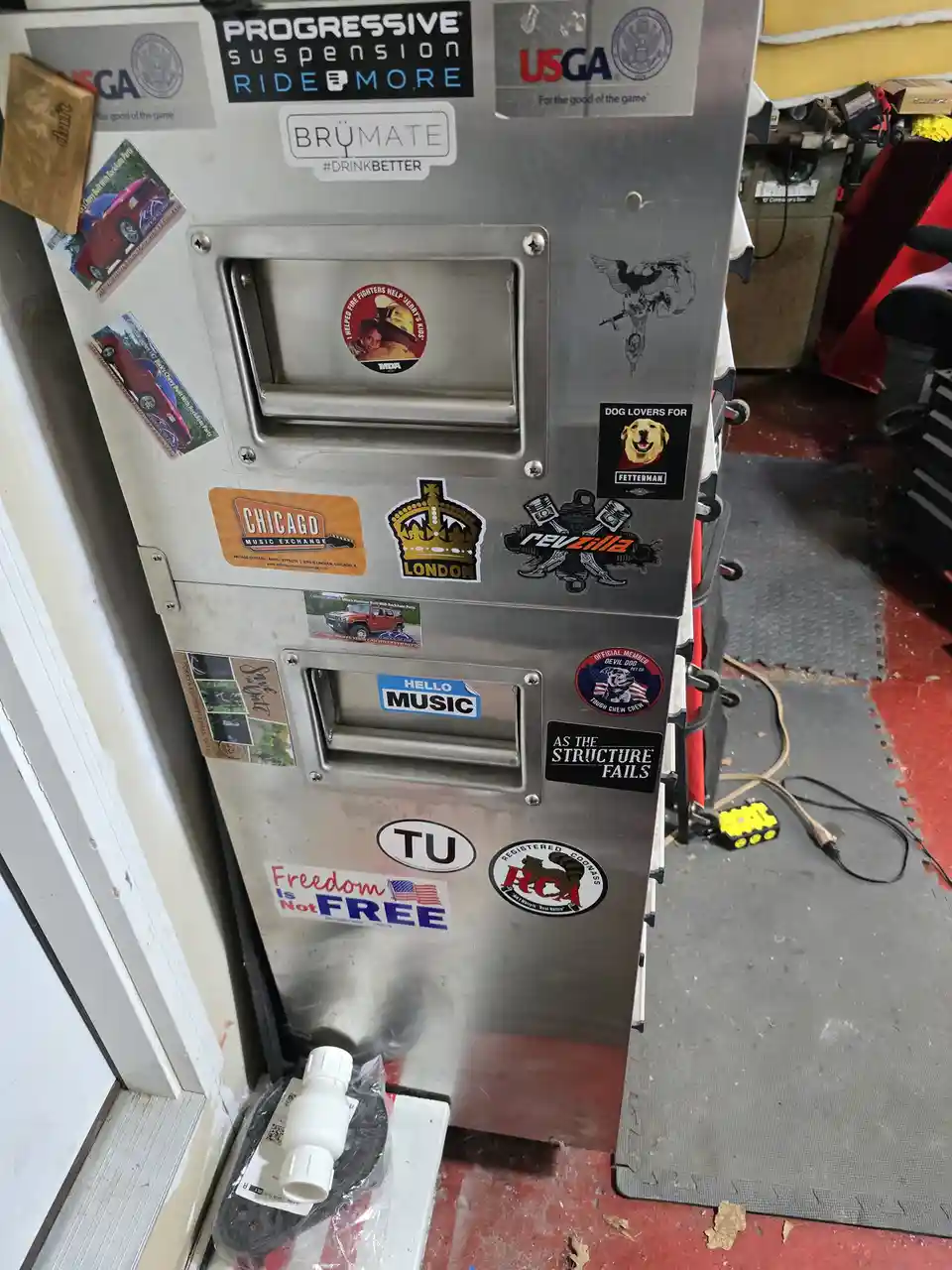 A stainless steel refrigerator covered in numerous stickers, including 'Progressive Suspension', 'USGA', 'BrüMate', 'Chicago…