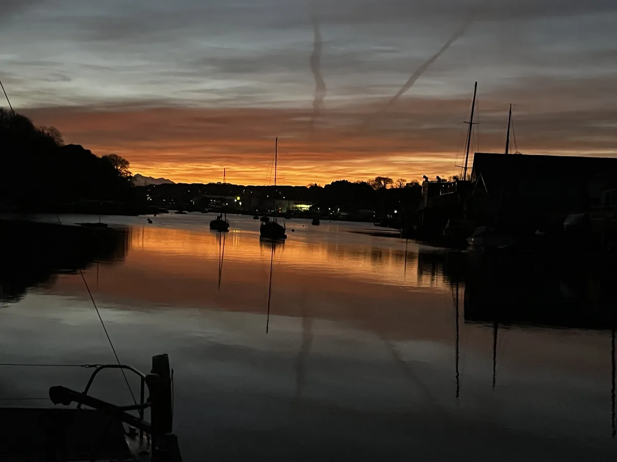 A serene harbor at dusk with boats silhouetted against a vibrant orange and gray sky, their masts reflected in calm water…