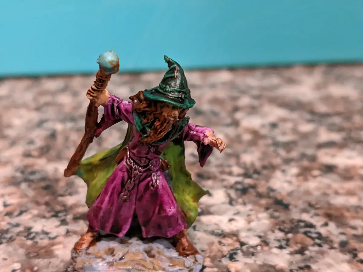 A painted miniature figurine of a wizard with a green pointed hat, purple robe, and green cape, holding a staff with a blue…