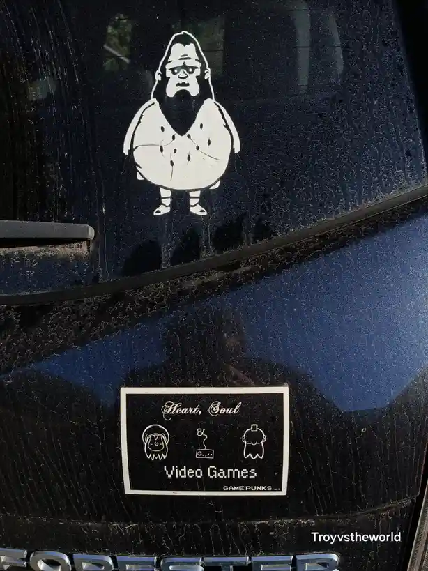 A close-up of a black car window with two white stickers. The top sticker is a cartoonish, overweight, bald man with a beard…