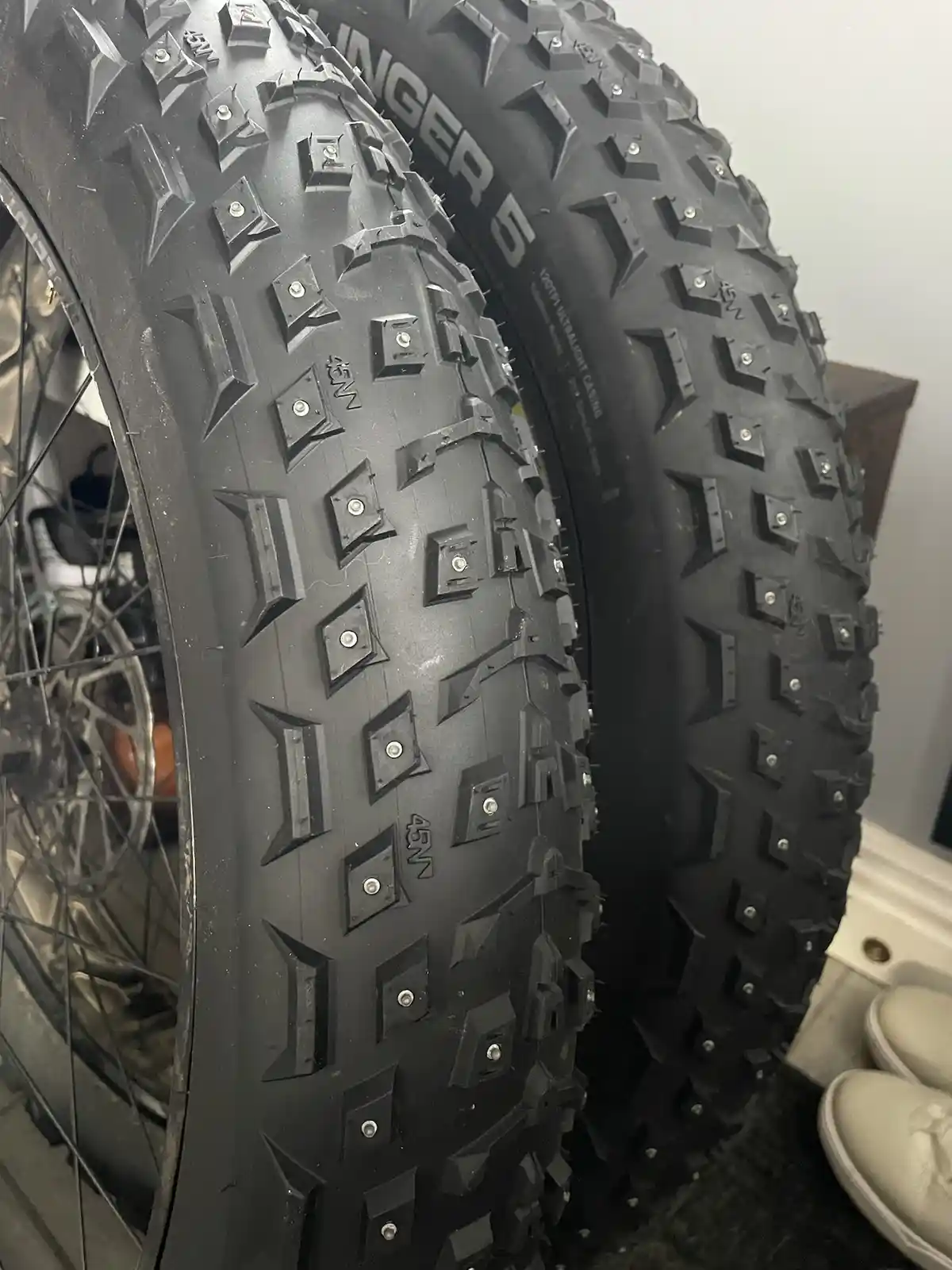 A close-up photo shows two large, black, knobby bicycle tires with aggressive tread patterns and visible metal studs…