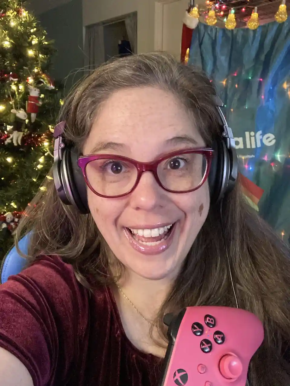A woman with glasses and headphones smiles widely while holding a pink Xbox controller, with a decorated Christmas tree and…