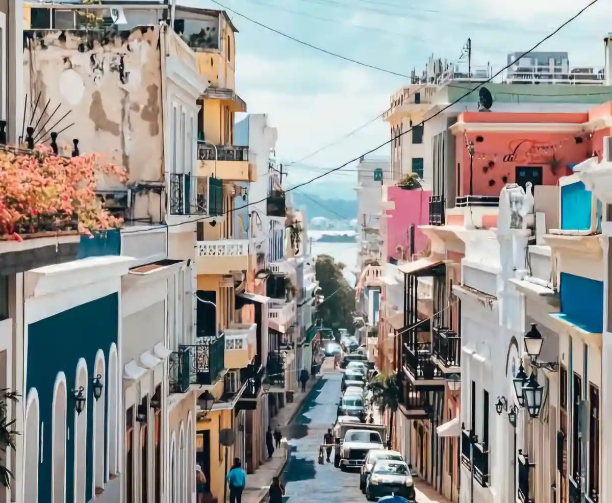 A vibrant, sunlit photo of a narrow, steep street in a colorful coastal town, flanked by multi-story buildings with…