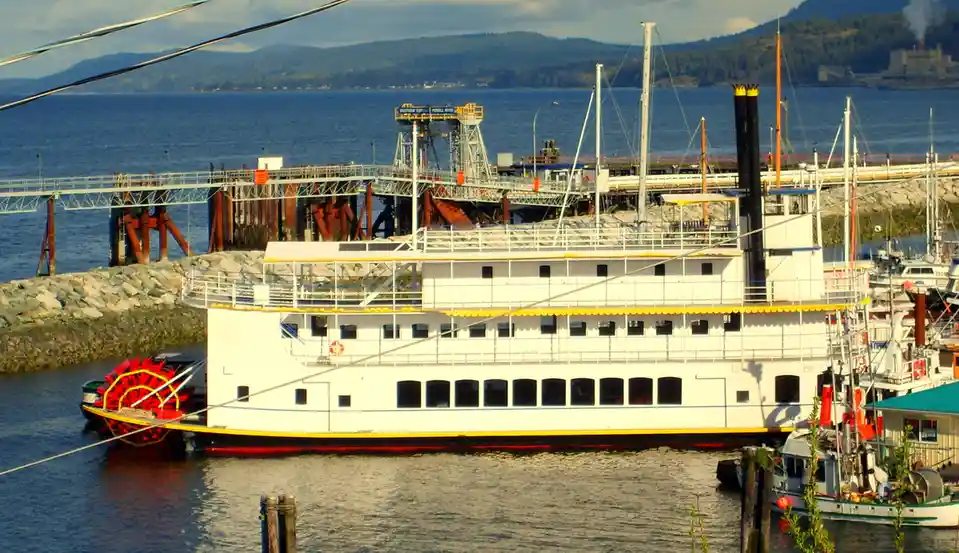 A large white paddlewheel steamboat with yellow trim is docked in a harbor, its red paddlewheel visible at the stern. Behind…