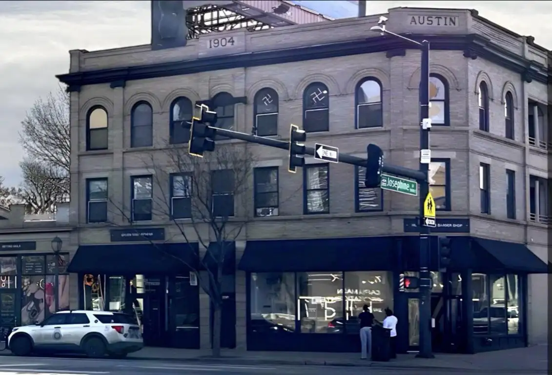 A street-level photo of a historic brick building at the corner of Austin and Josephine streets, with a silver police SUV…