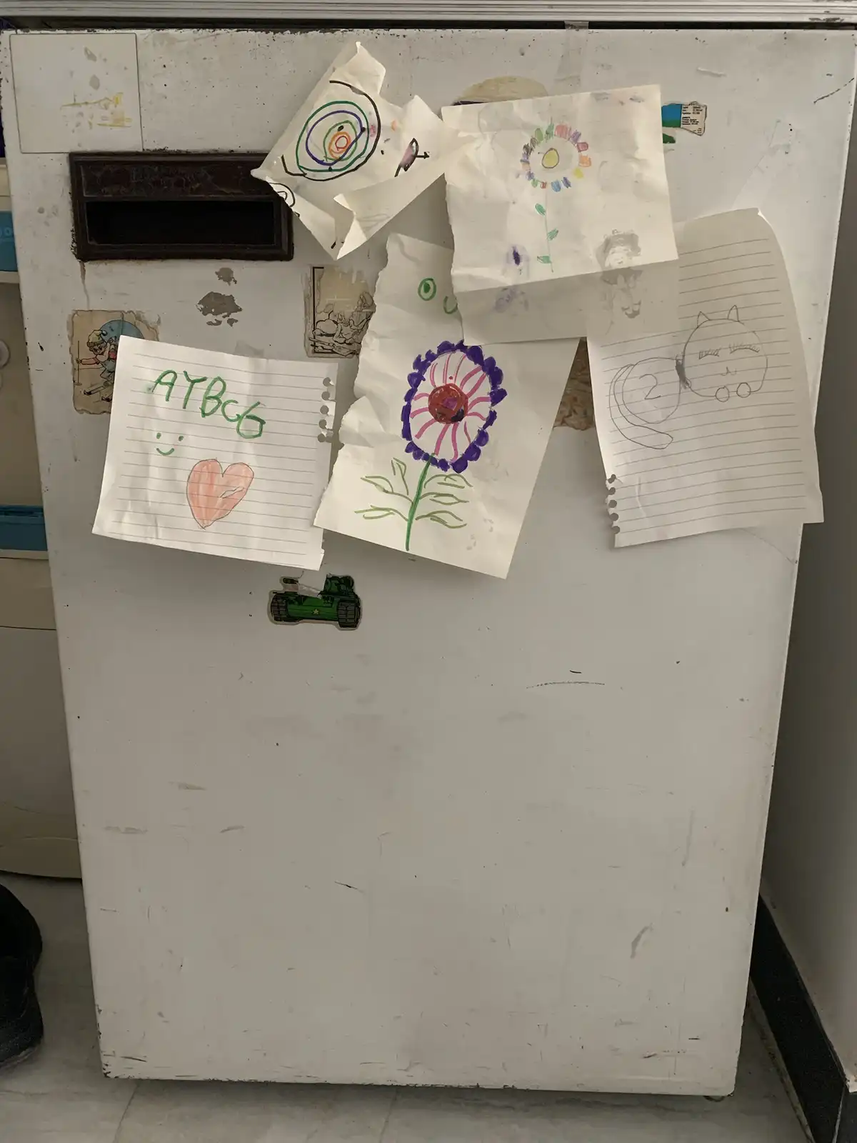 A white refrigerator door covered in various child's drawings on lined paper, including a flower, a cat, and abstract…