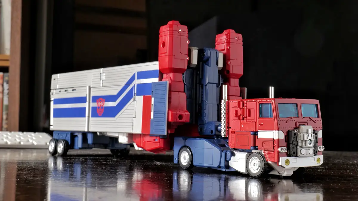 A red, blue, and silver toy truck with a trailer, featuring the Autobot logo on its side, is shown in a detailed close-up on…