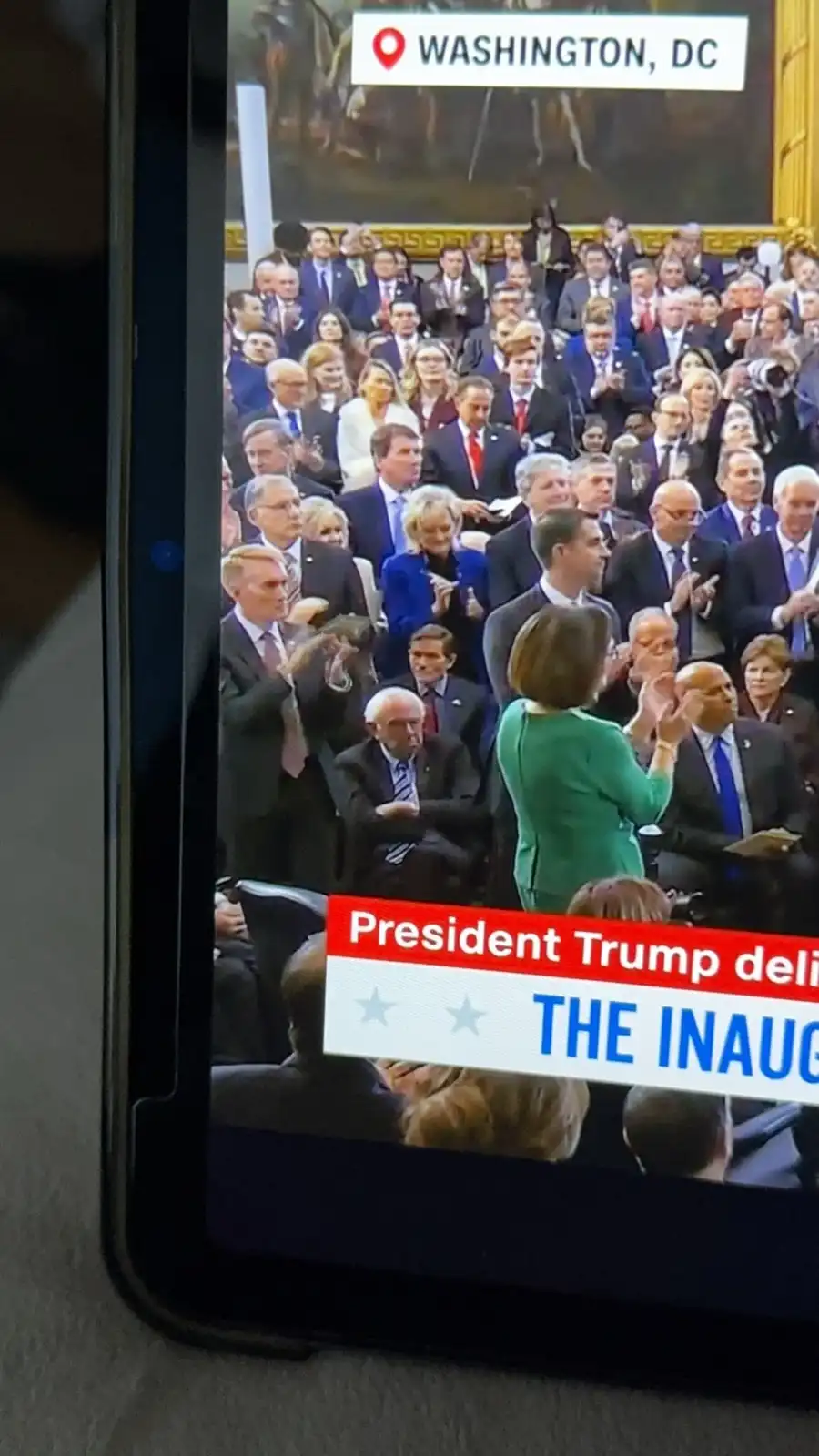 A smartphone screen displays a news broadcast from Washington, DC, showing a crowd of formally dressed people applauding…