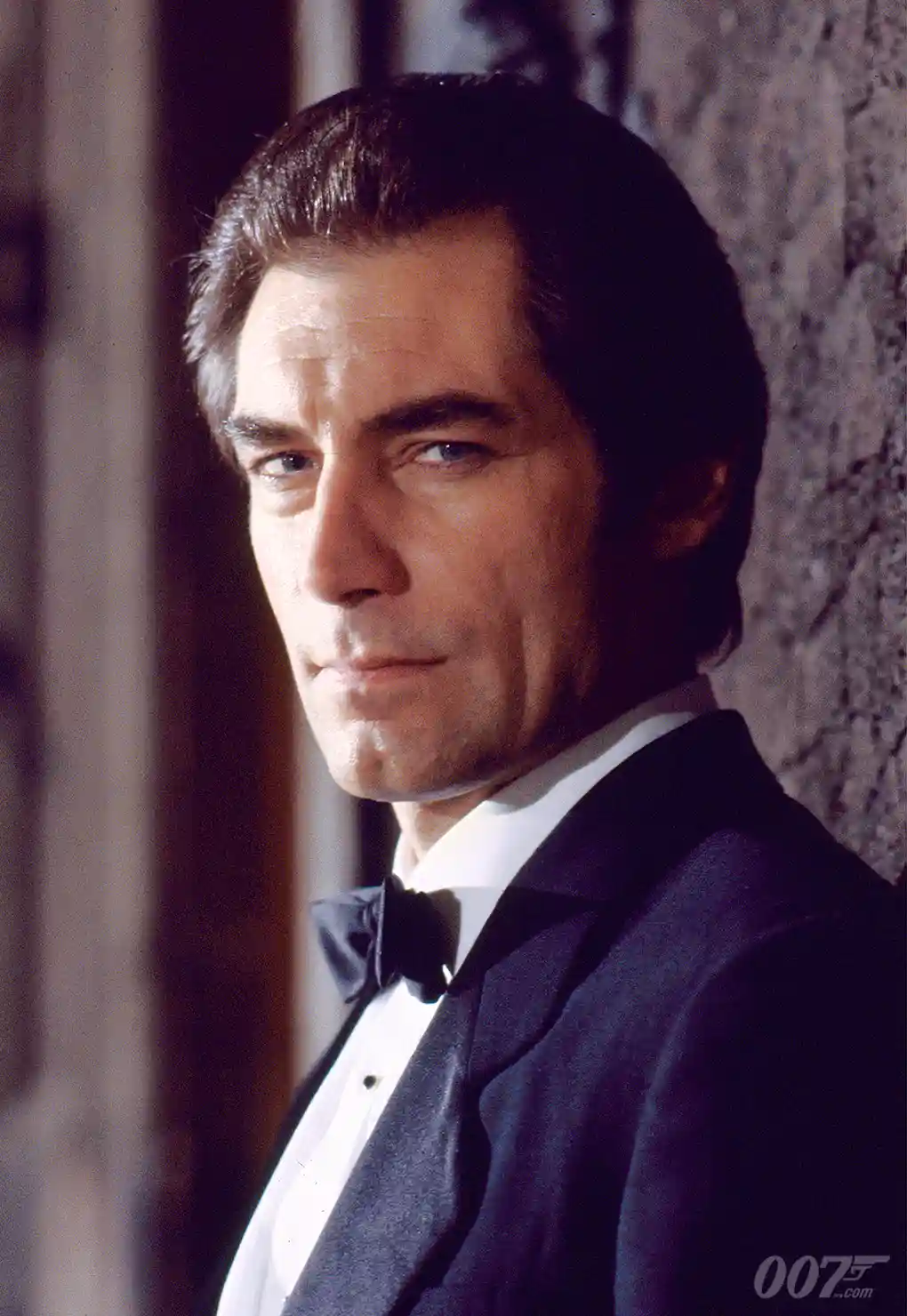 a still image of actor Timothy Dalton as James Bond, dressed in a dark tuxedo with a bow tie, looking over his shoulder with…
