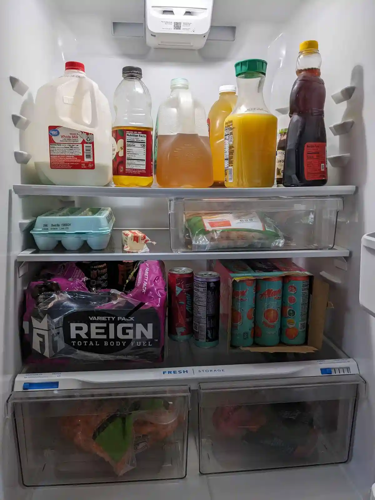 A refrigerator interior filled with beverages and food, prominently featuring a large 'REIGN TOTAL BODY FUEL' energy drink…
