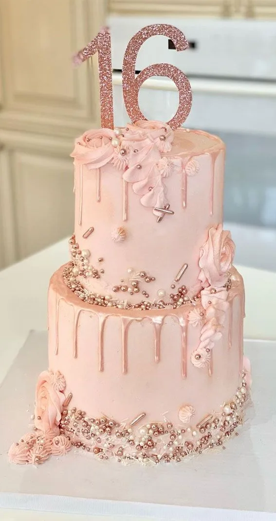 A two-tiered pink drip cake decorated with sugar flowers, pearls, and rose gold accents, topped with a glittery number 16…