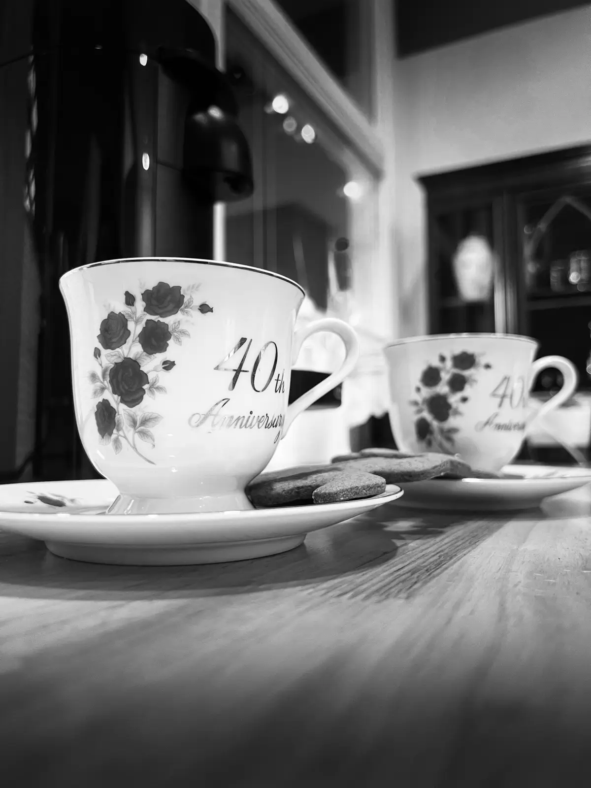 A black-and-white photo shows two matching porcelain teacups with floral patterns and the text '40th Anniversary' on them…