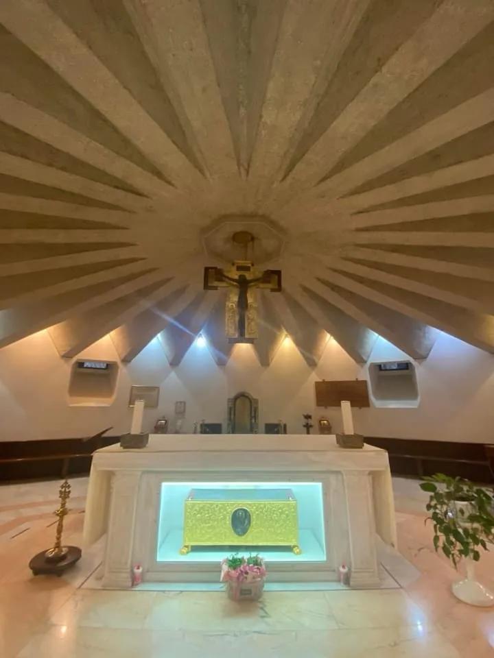 A photo of an ornate church altar with a golden reliquary under a radiant, sunburst-style concrete ceiling. A crucifix hangs…
