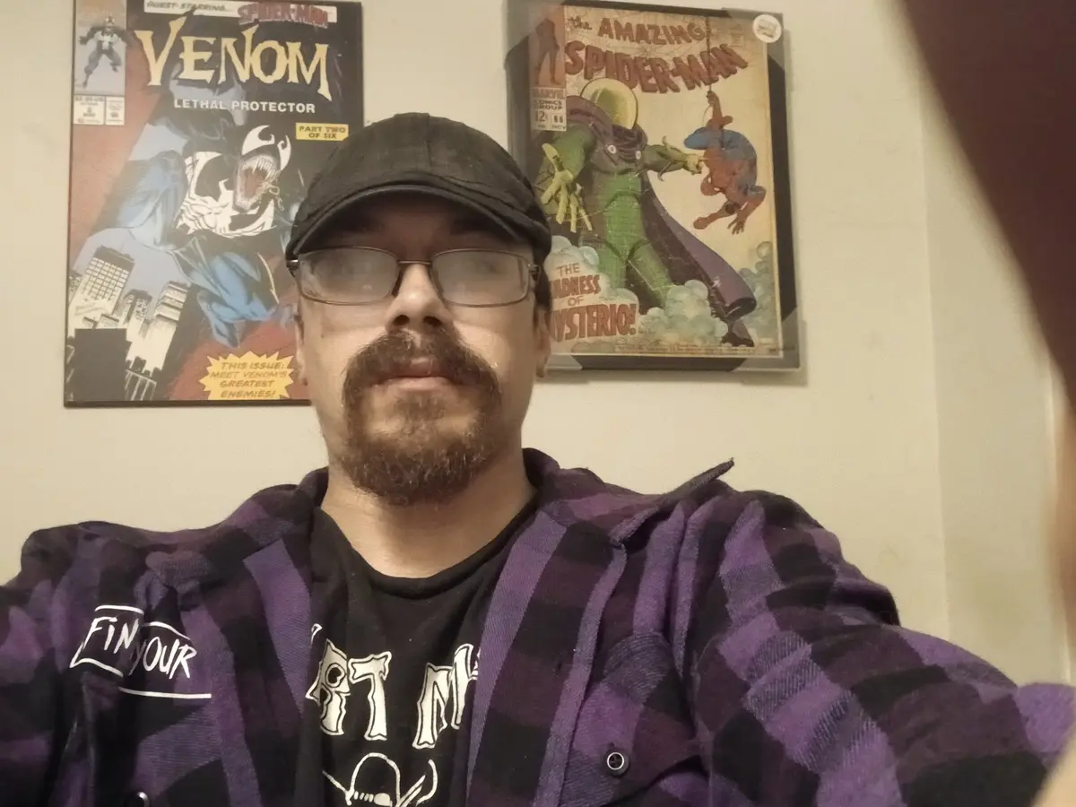 A man with glasses and a mustache, wearing a purple plaid shirt over a black t-shirt, takes a selfie. Behind him on the wall…