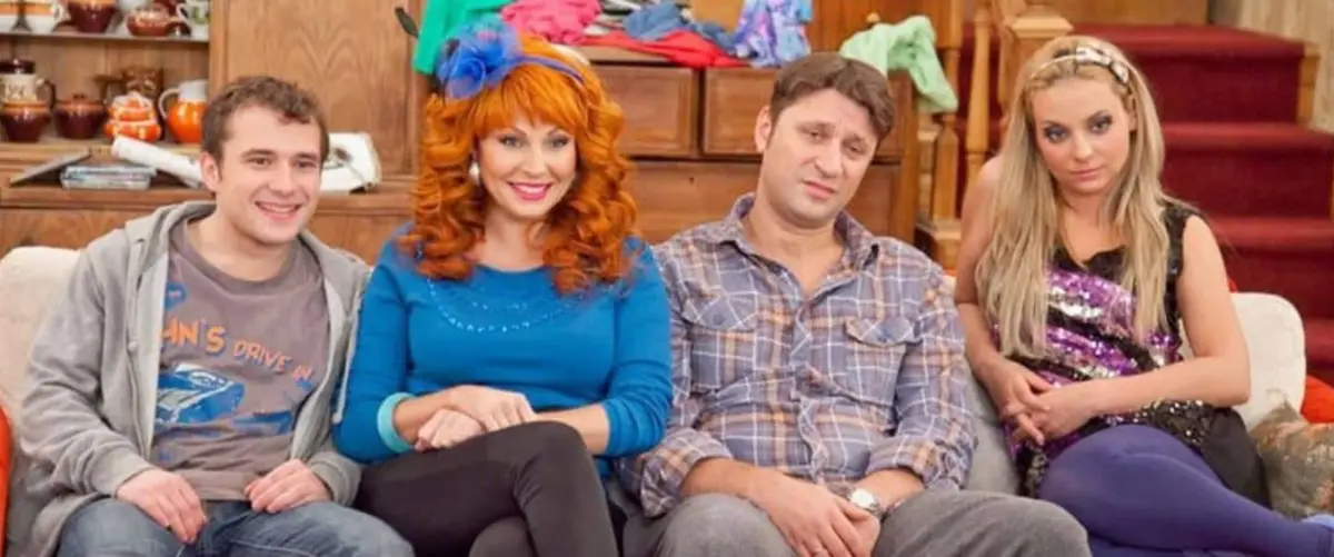 Four people sit on a couch in a cluttered living room; the man on the far left smiles brightly, the woman next to him with…