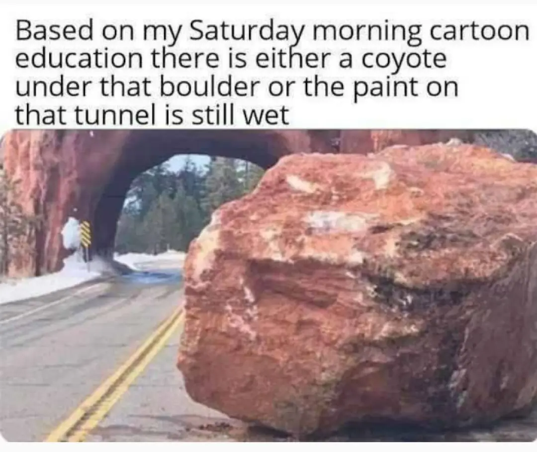 A large reddish-brown boulder blocks a paved road with yellow double lines, directly in front of a tunnel carved into a…