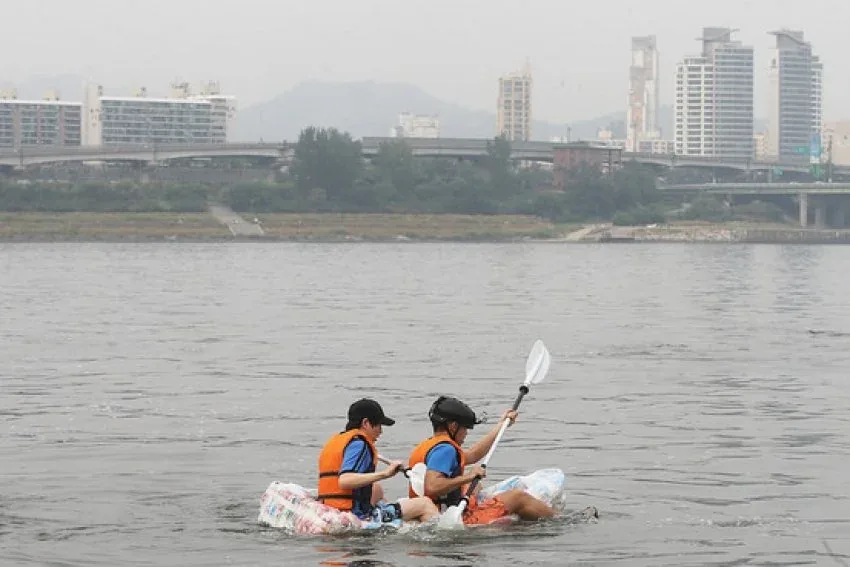Two people are paddling a makeshift raft made of plastic bottles on a river, wearing life vests and helmets, with a city…