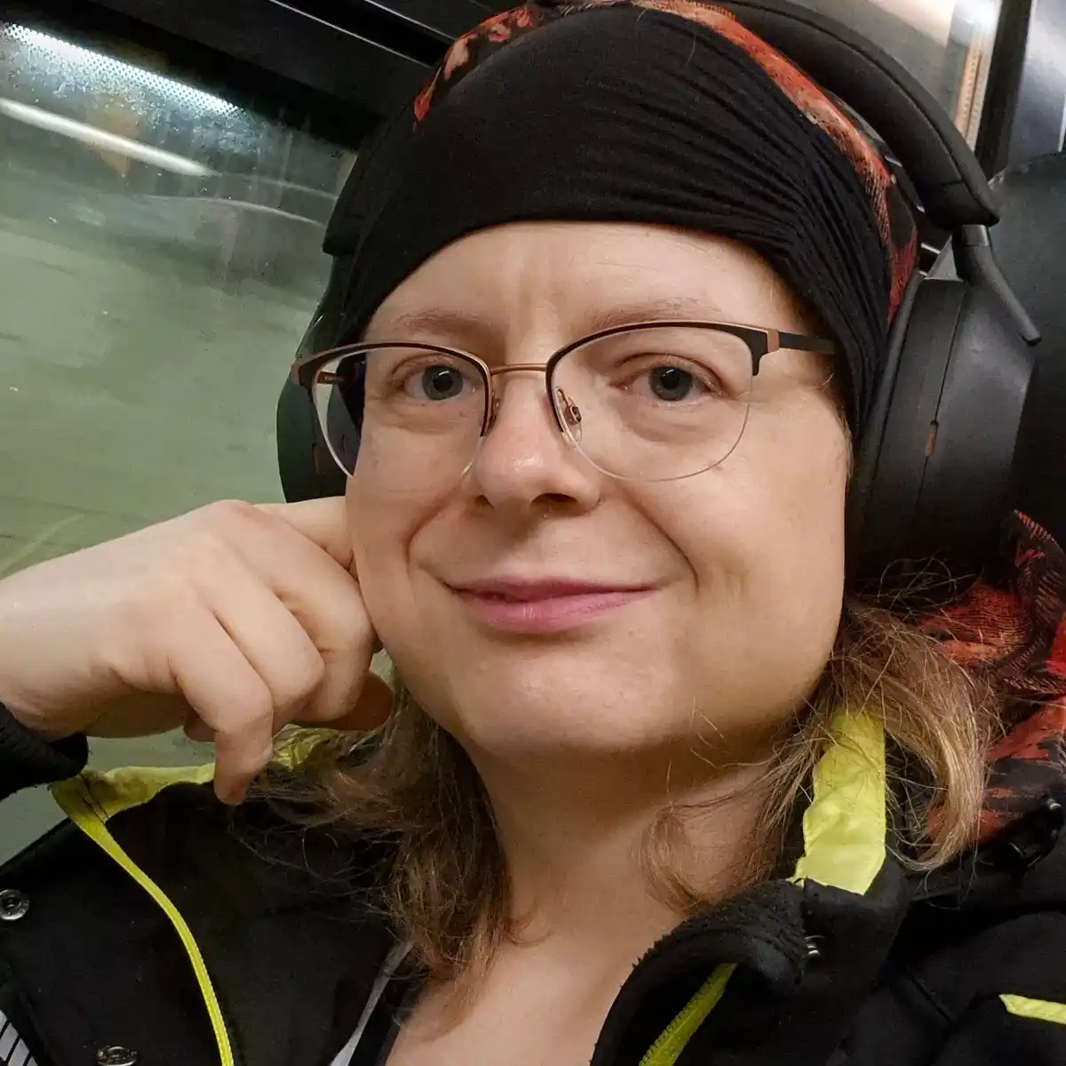 A person wearing glasses, a black headscarf with red-orange patterns, and large black headphones, smiles while resting their…