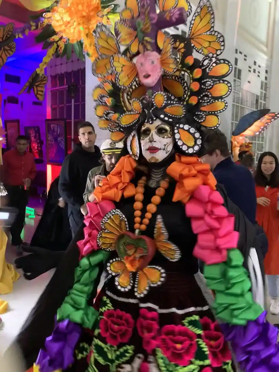 A person in a vibrant, elaborate Day of the Dead costume with a large butterfly headdress, sugar skull makeup, and colorful…