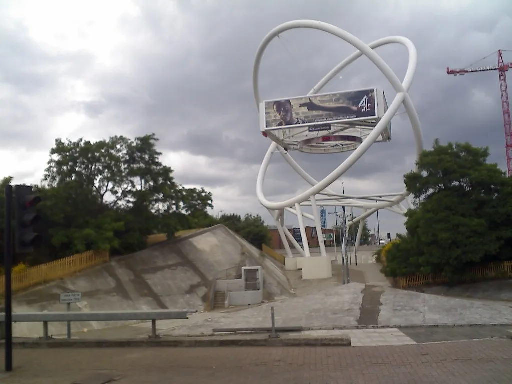 An outdoor photo shows a large white sculpture resembling an atom with a billboard inside it. The billboard displays a man…