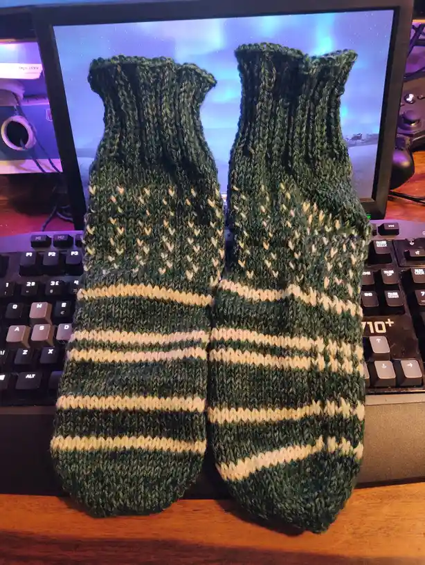 A pair of handmade green and cream knitted socks with a heart pattern and striped design are displayed upright on a wooden…