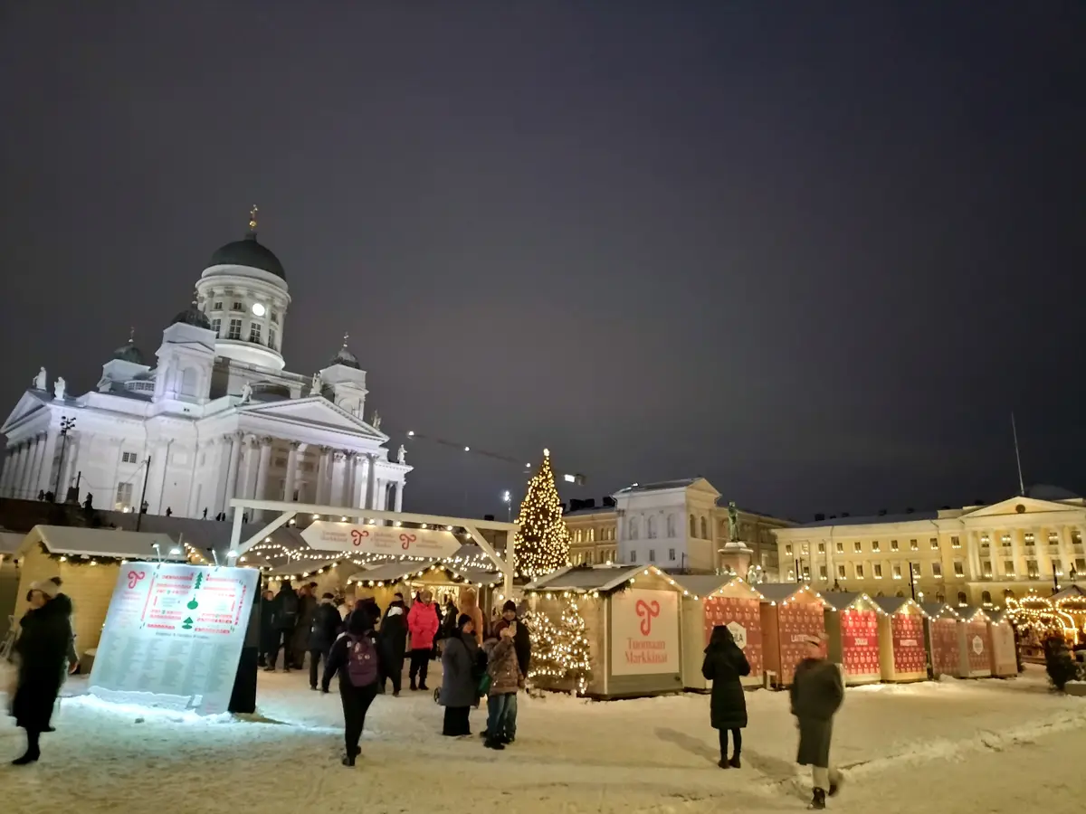 A nighttime photo of a snowy Christmas market in front of a large, illuminated white neoclassical cathedral with domes…