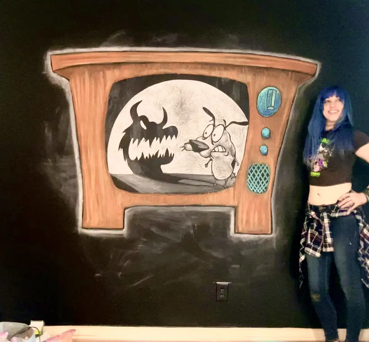 A person with blue hair stands beside a chalk drawing of a vintage TV on a blackboard. The TV screen shows a cartoon scene…