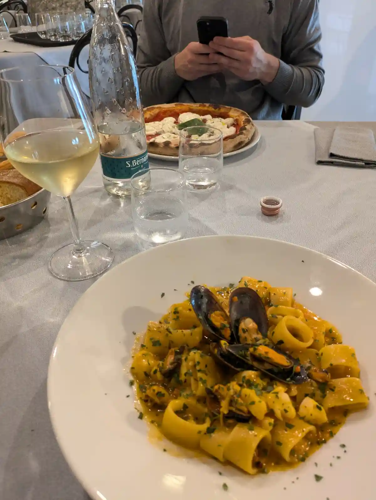 A person in a grey sweater sits at a restaurant table, focused on their phone, while a plate of pasta with mussels and a…