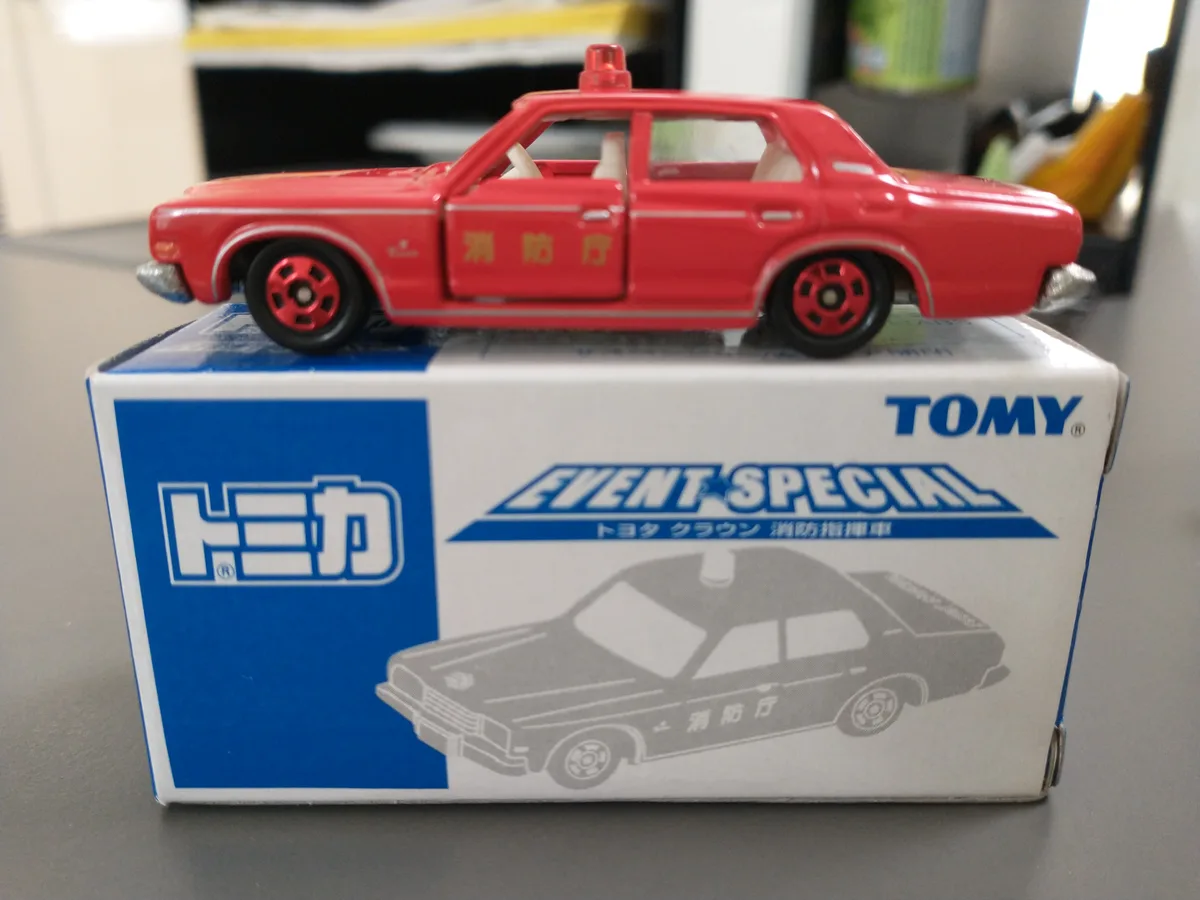 A red toy car with '消防厅' written on its side sits atop its original Tomy 'Event Special' box, which features an illustration…