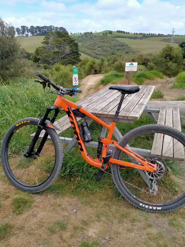 An orange mountain bike is leaning against a wooden picnic table in a grassy, hilly outdoor area. Behind the bike, a sign…