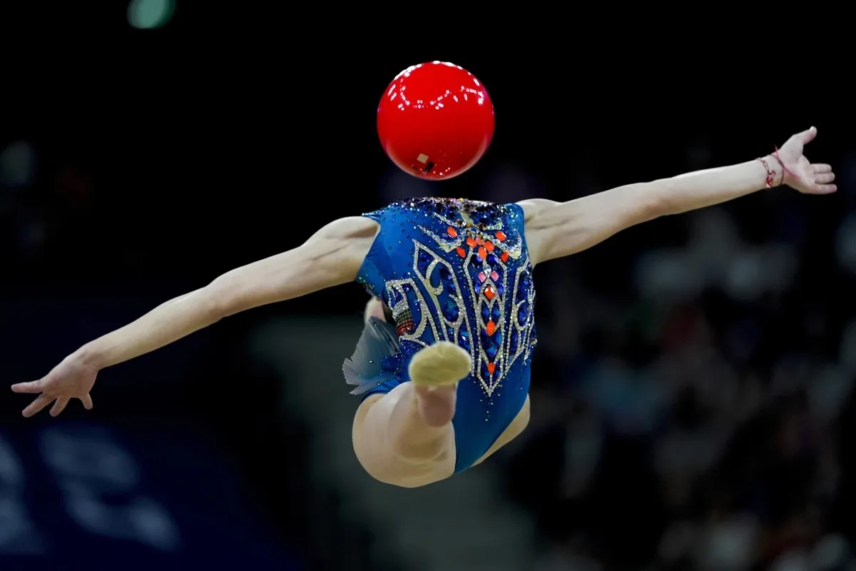 A female rhythmic gymnast in a blue, bejeweled leotard is captured mid-air during a dynamic pose, her arms extended and one…