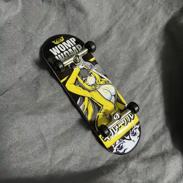 A small fingerboard skateboard with a black and yellow anime girl graphic, featuring the text 'WOMP WOMP' in bold white…