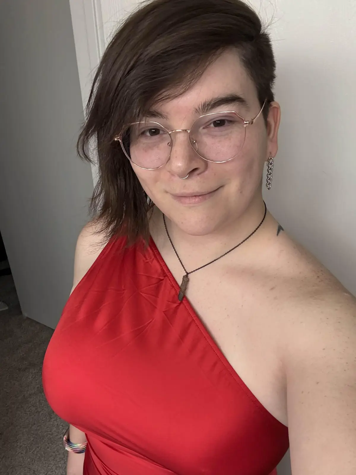 A person with short brown hair, wearing rose gold glasses, a red one-shoulder dress, a nose ring, and chain earrings, smiles…