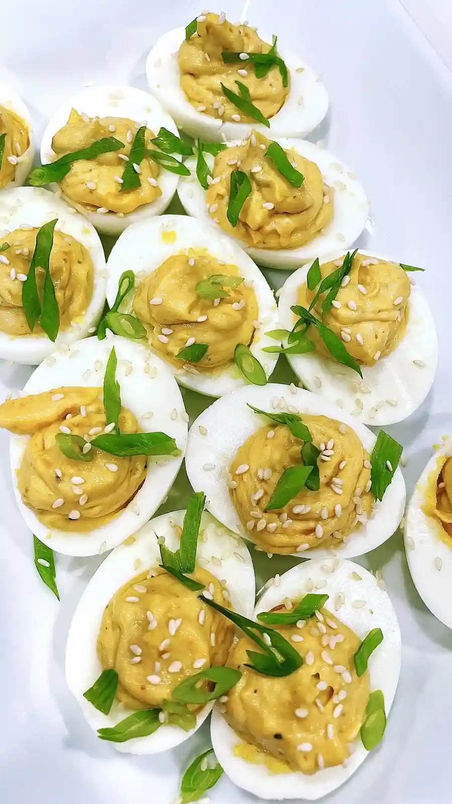 A close-up photo shows a white plate filled with neatly arranged deviled eggs. Each egg half is filled with a creamy yellow…