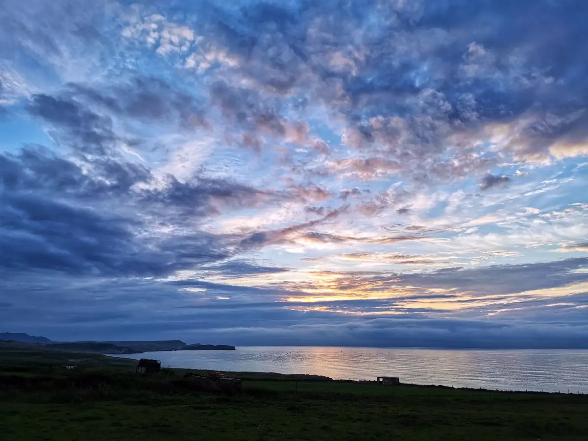 A scenic photo of a coastal landscape at dawn or dusk, featuring a vast sky filled with dramatic blue and pink clouds over a…