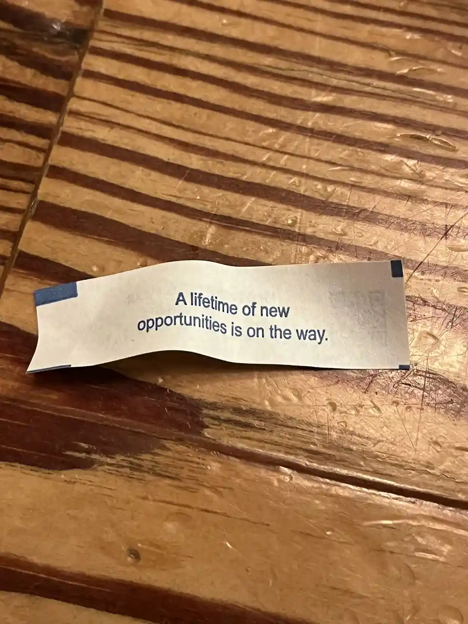 A crumpled fortune cookie slip with the text 'A lifetime of new opportunities is on the way.' lies on a worn wooden surface…