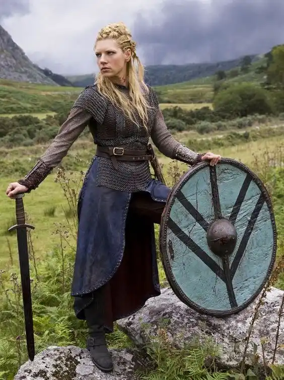 A woman in Viking armor stands outdoors holding a sword and shield, looking sternly at the camera. The overlaid text reads…