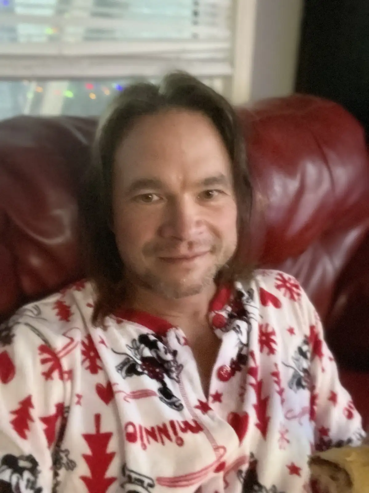 A man with shoulder-length brown hair sits in a red leather chair wearing festive white pajamas with red snowflakes, hearts…