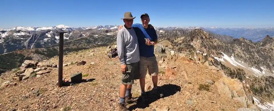 Two smiling men pose for a photo on a rocky mountain summit with a vast, snow-capped mountain range in the background under…
