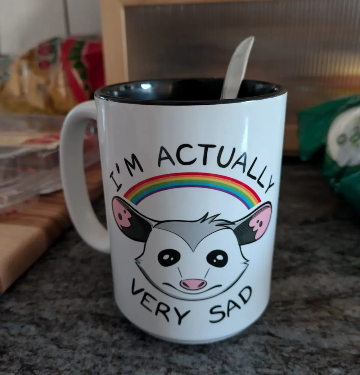 A white ceramic mug with a black rim sits on a kitchen counter, featuring a cartoon illustration of a sad-faced opossum with…