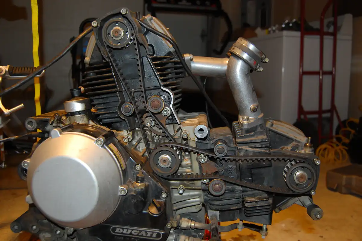 A close-up photo of a Ducati motorcycle engine with its timing belt and gears exposed, sitting in a garage. The overlaid…