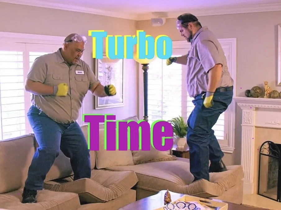 Two men in work clothes and safety goggles stand on opposite ends of a beige couch in a living room, appearing to be in a…