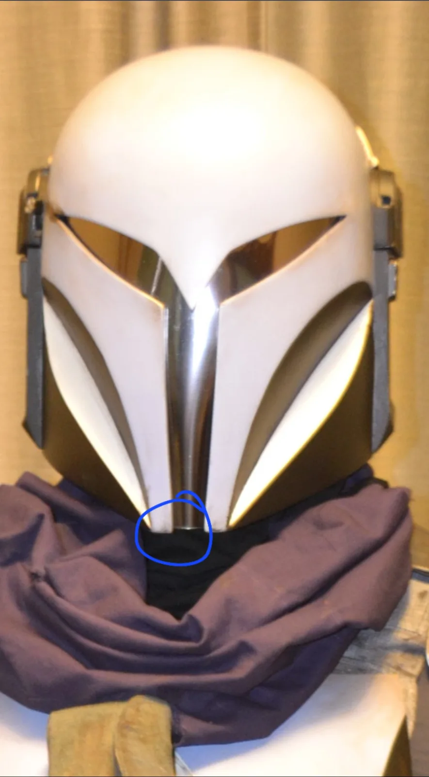 A white and gold Mandalorian-style helmet, likely a cosplay prop, is displayed on a stand with a purple cloth draped around…